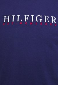 Námorně modré bavlněné tričko s bílým vyšitým textem "HILFIGER" a červenými akcenty s "EST. MCMLXXV" pod ním. Hladká textura, design s kulatým výstřihem.