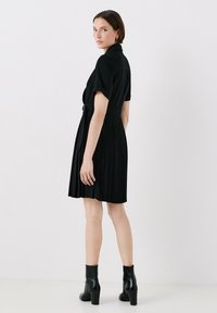 Robe plissée noire avec des manches courtes et une taille ceinturée. Elle présente un col et arrive juste au-dessus des genoux. Portée avec des bottes à talons noires.