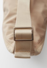 Borsa beige con una superficie in tessuto texturizzato e una larga tracolla intrecciata, dotata di una fibbia in metallo lucido per la regolazione della lunghezza.