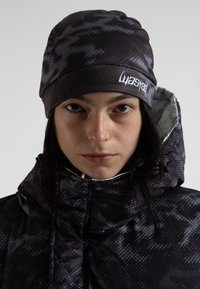 Gorro negro con un patrón, de textura suave, con un logo. Debajo, una chaqueta acolchada con un diseño geométrico oscuro y cuello alto.