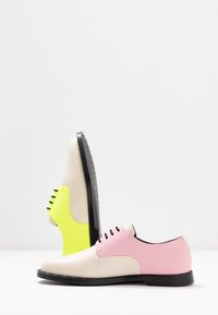 Chaussures habillées en cuir bicolore avec semelles et lacets noirs ; une chaussure rose et crème, l'autre jaune fluo et crème, présentées sur une surface blanche.