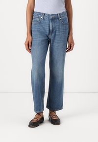 Jeans de denim azul claro com um design de perna reta, cintura média e desbotamento sutil. Combinados com sapatilhas pretas com tiras cruzadas.