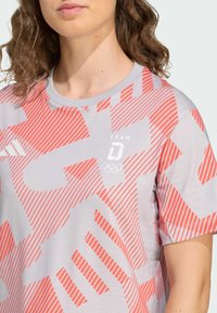 Szary T-shirt z wyrazistym czerwonym paskowanym wzorem, okrągłym dekoltem i logo olimpijskim. Wykonany z lekkiego, oddychającego materiału.