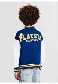 Granatowa kurtka varsity z białymi rękawami. Posiada żółte napisy "PLAYER" i "CALIFORNIA", grafikę lwa oraz paskowane mankiety.