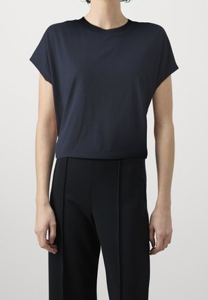 Kobieta w granatowej dopasowanej bluzce z krótkim rękawem, wpuszczonej w czarne, dopasowane, wysokowłosowe spodnie, stojąca na jednolitym tle.