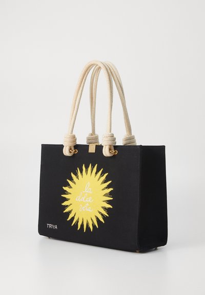 Black canvas tote bag with cream rope handles and gold hardware. Features a yellow sun graphic with "la dolce vita" text. Brand name TRIYA.