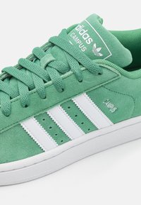 Vihreät keinonahkaisista Adidas Campus -tennarit, joissa on valkoiset raidat, valkoinen pohja ja yhteensopivat vihreät nauhat valkoista taustaa vasten.