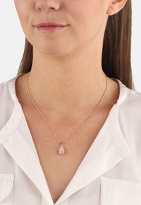 Collar de oro rosado con una delicada cadena y un colgante en forma de pera que presenta múltiples pequeñas piedras brillantes. Llevado sobre una blusa blanca.