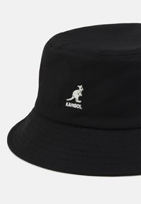 Gorro de cubo negro hecho de tejido suave, con un logo de Kangol que presenta un contorno de canguro blanco en la parte frontal. Textura sutil y estriada en el ala.
