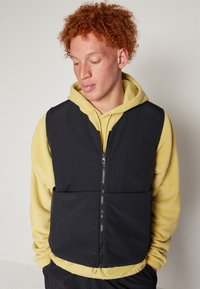 Μαύρο αμάνικο γιλέκο με φερμουάρ μπροστά, υφή υφάσματος, δύο μπροστινές τσέπες, φορεμένο πάνω από μια κίτρινη φούτερ με κορδόνι και ελαστικές μανσέτες.