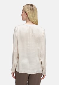 Beige Bluse mit langen Ärmeln, einer glatten Textur, einem gefältelten Rückendetail und geknöpften Manschetten, getragen mit braunen Hosen.
