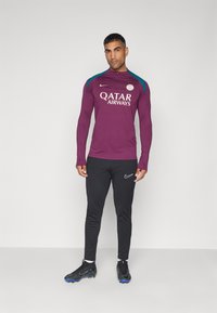 Musta pitkähihainen harjoituspaita turkooseilla korostuksilla, jossa on "Qatar Airways" -logo, yhdistettynä mustiin Nike -housuihin ja kengissä.