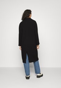 Mujer con cabello rizado, vistiendo un abrigo largo negro, jeans azules y zapatillas blancas, de pie contra un fondo blanco liso, mirando hacia atrás.