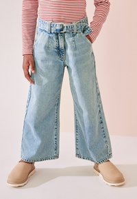 Ljusblå denimjeans med vida ben, bälte och framfällor, i kombination med beige slip-on skor, visade mot en neutral bakgrund.