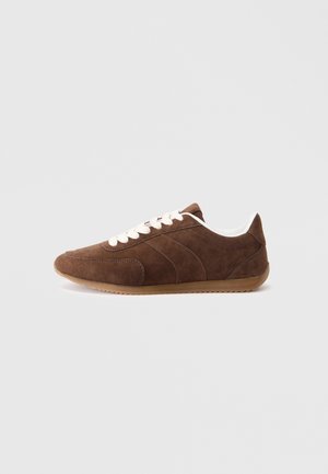 Brązowy zamszowy sneaker z białymi sznurowadłami, o niskim profilu i gumowej podeszwie, pokazany z boku na jasnoszarym tle.
