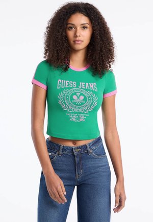Zelené skrátené tričko s ružovým lemovaním, na prednej strane s grafikou a emblémom "GUESS JEANS". Skombinované s modrými džínsami s vysokým pásom.