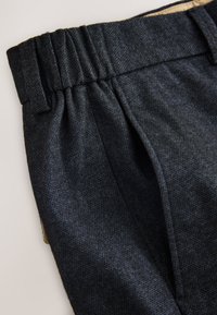 Pantalon en tissu bleu foncé texturé avec une taille froncée et des poches latérales. Le matériau semble tissé avec un motif subtil.