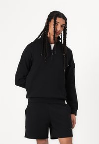 Zwarte zacht fleece pullover met een ritssluiting bij de kraag, gecombineerd met zwarte shorts. Beide kledingstukken hebben een zachte textuur en een minimalistisch ontwerp.