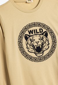Beige sweatshirt med en svart tiger grafik och ordet "WILD" inuti en cirkulär design med ett geometriskt mönster.