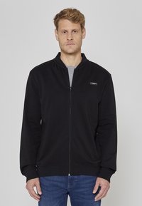 Veste noire à fermeture éclair fabriquée en tissu doux, avec un col montant, des manches longues et un petit logo sur le côté gauche de la poitrine. Portée par-dessus une chemise grise.