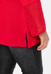 Blouse rouge avec un corps en tissu uni, des manches transparentes à pois, des fentes latérales et une coupe décontractée, associée à un denim foncé.