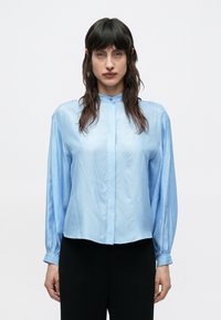 Žena s dlhými tmavými vlasmi, oblečená v svetlomodrej hodvábnej blúzke s zapnutými gombíkmi na manžetách a čiernych nohaviciach, na bielom jednofarebnom pozadí.