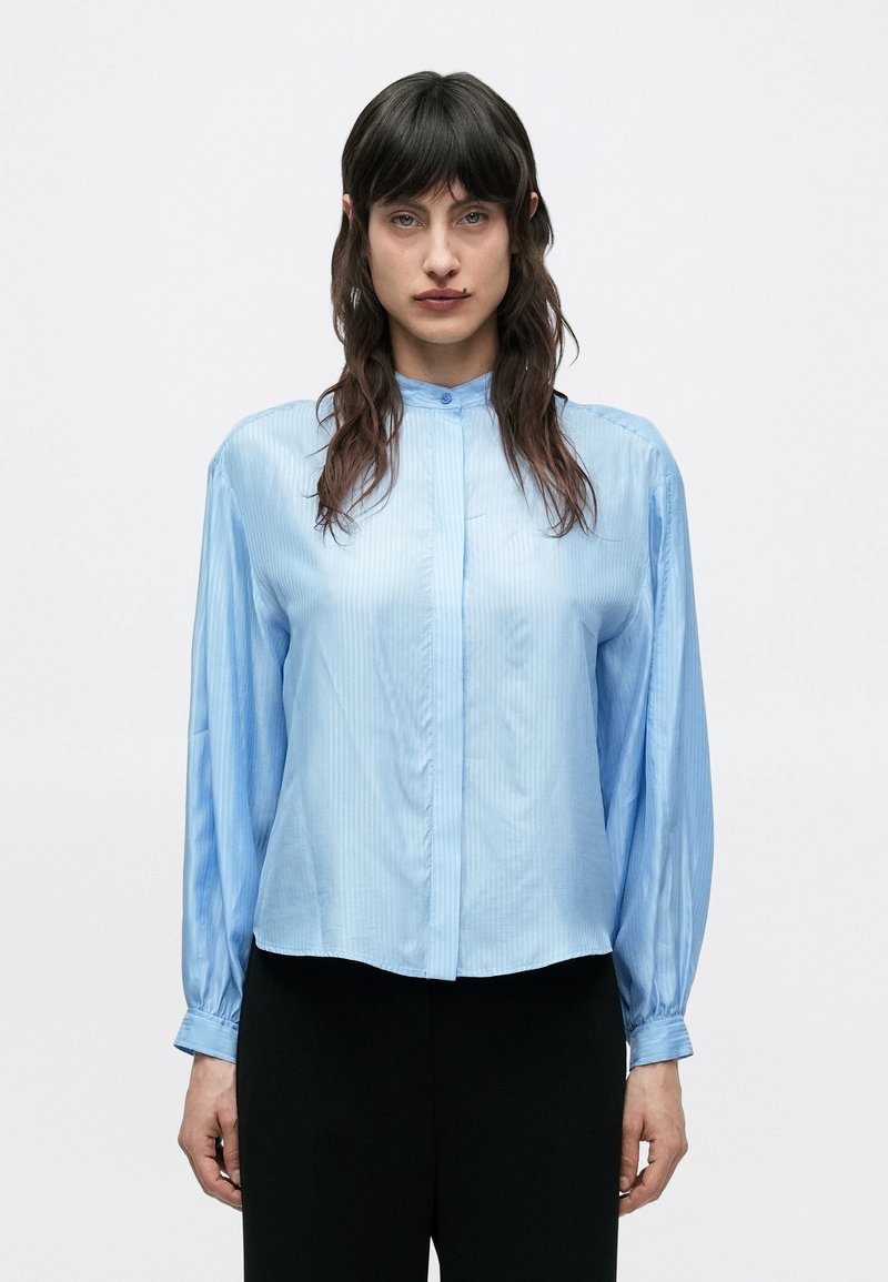 Žena s dlhými tmavými vlasmi, oblečená v svetlomodrej hodvábnej blúzke s zapnutými gombíkmi na manžetách a čiernych nohaviciach, na bielom jednofarebnom pozadí.