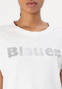 Camiseta de algodón blanca con un escote amplio y mangas cortas, adornada con letras de tachuelas plateadas que dicen "Blauer."