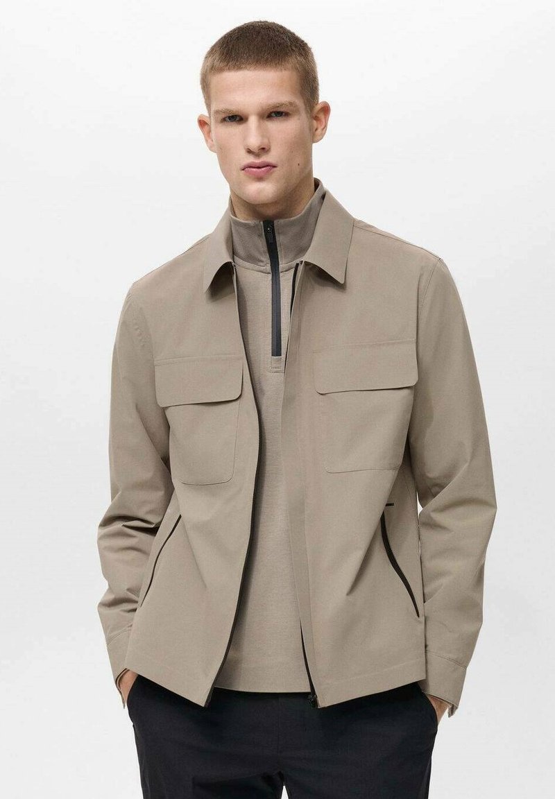 Ljusbeige jacka med en framficklås, två bröstfickor, en hög krage och en slät textur. Styld över en matchande topp.