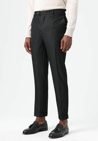 Schwarze, maßgeschneiderte Hose mit strukturierter Oberfläche, schmaler Passform, vorderseitigen Bundfalten und umgeschlagenen Säumen. Kombiniert mit schwarzen Loafers.