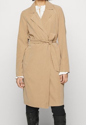 Frau trägt einen knieumspielenden beigen Wickelmantel mit Taillenschärpe über einer weißen Bluse und schwarzen Hosen sowie schwarzen kniehohen Stiefeln.