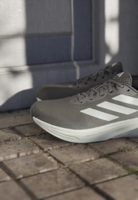 Szare buty sportowe z teksturowanym siateczkowym wierzchem, białym wzorem z trzema paskami i smukłą sylwetką, ustawione na kamiennej powierzchni.