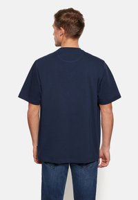 Tricou din bumbac de culoare bleumarin, cu mâneci scurte și decolteu rotund. Textură netedă, croială lejeră și tivul din spate ușor mai lung.