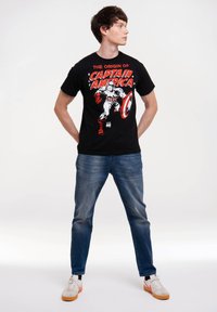 Schwarzes Baumwoll-T-Shirt mit einem Grafikdesign von Captain America, mit roten und weißen Akzenten. Tragen mit blauen Jeans und weißen Schuhen.