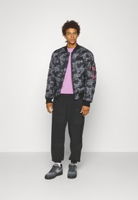 Černá maskovací péřová bunda s žebrovanými manžetami, zkombinovaná se světle fialovým svetrem a černými cargo kalhotami. Šedé tenisky dokončují vzhled.