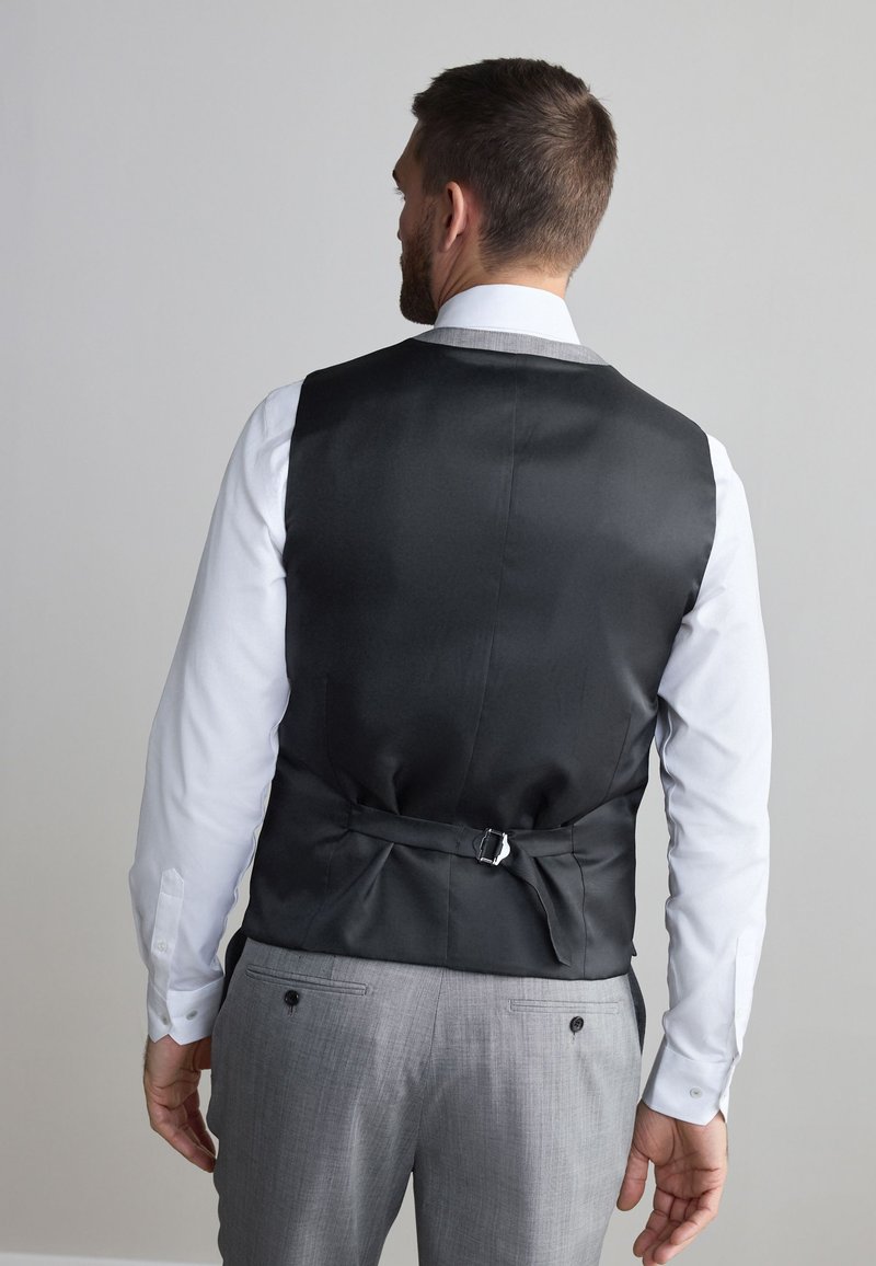Homme portant un gilet noir par-dessus une chemise blanche et un pantalon gris, vu de dos sur un fond clair uni.