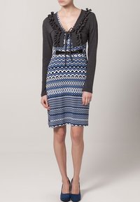 Sukienka do kolan z ciemnoszarym, falbaniastym kardiganem na wierzchu oraz spódnicą w niebiesko-białą zygzakowatą wzór. Ma wiązanie w talii.