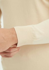 Pull en maille beige avec un poignet côtelé, texture lisse et coupe ajustée. Gros plan sur le bras en train de tirer la manche.