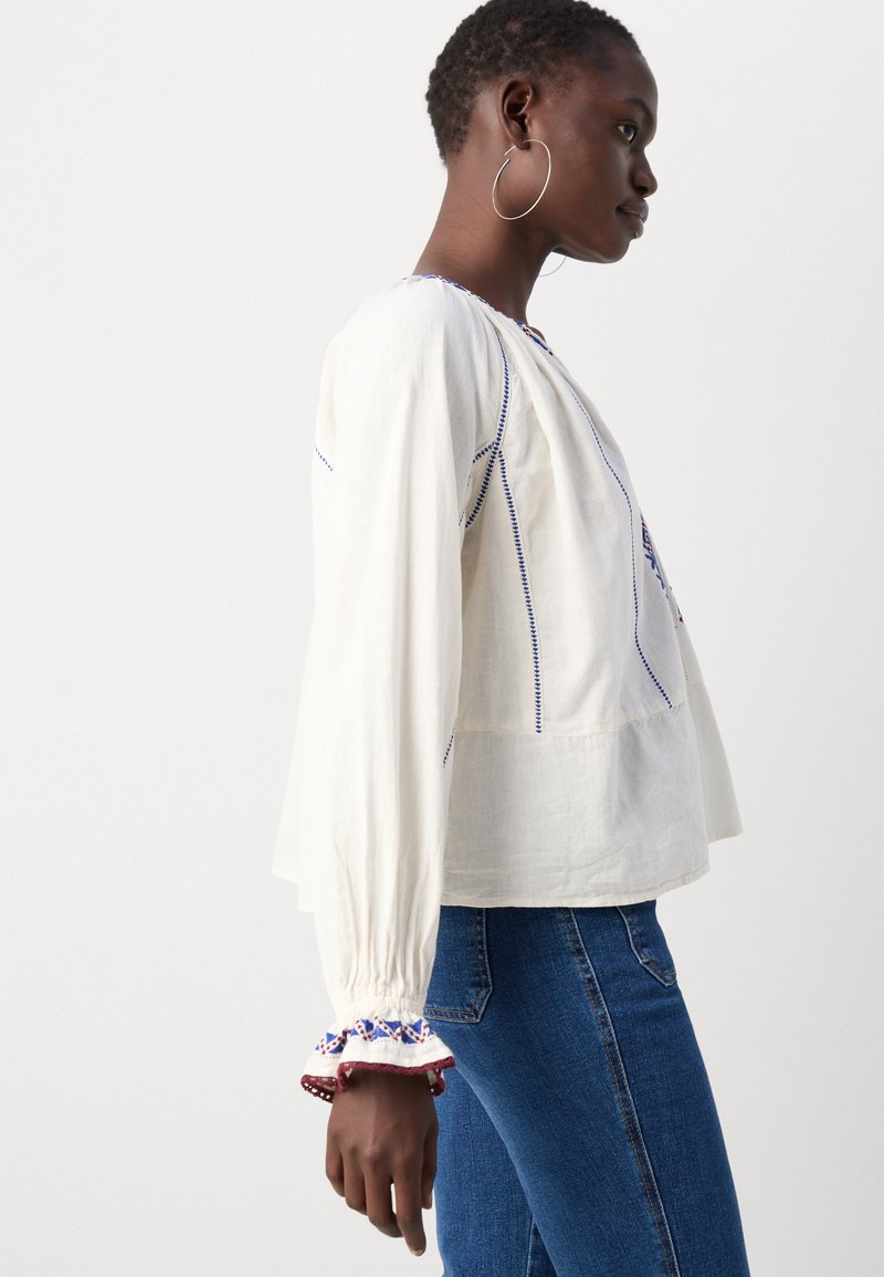 Blouse blanche à manches longues avec des accents de broderie bleus, une coupe ample et des poignets froncés. Associée à un jean en denim.