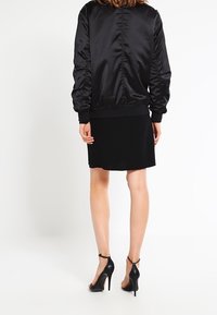 Svart satin bomberjacka med ribbad krage och manschetter, bärs över en åtsittande svart klänning, i kombination med svarta högklackade sandaler.