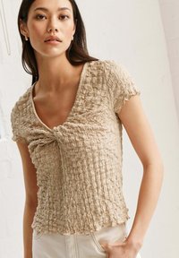 Haut en tissu beige froissé à manches courtes, doté d'un décolleté en V peu profond et d'un détail en torsion à l'avant. Porté avec un pantalon de couleur claire.
