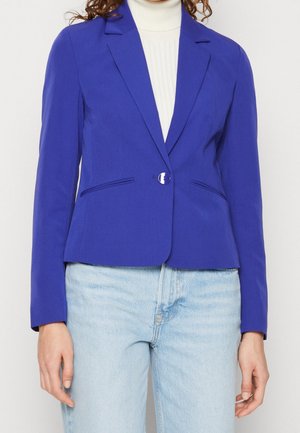 Femme portant un blazer bleu vif cintré avec un bouton sur un col roulé blanc et un jean bleu clair, montrant le haut du torse et les bras.