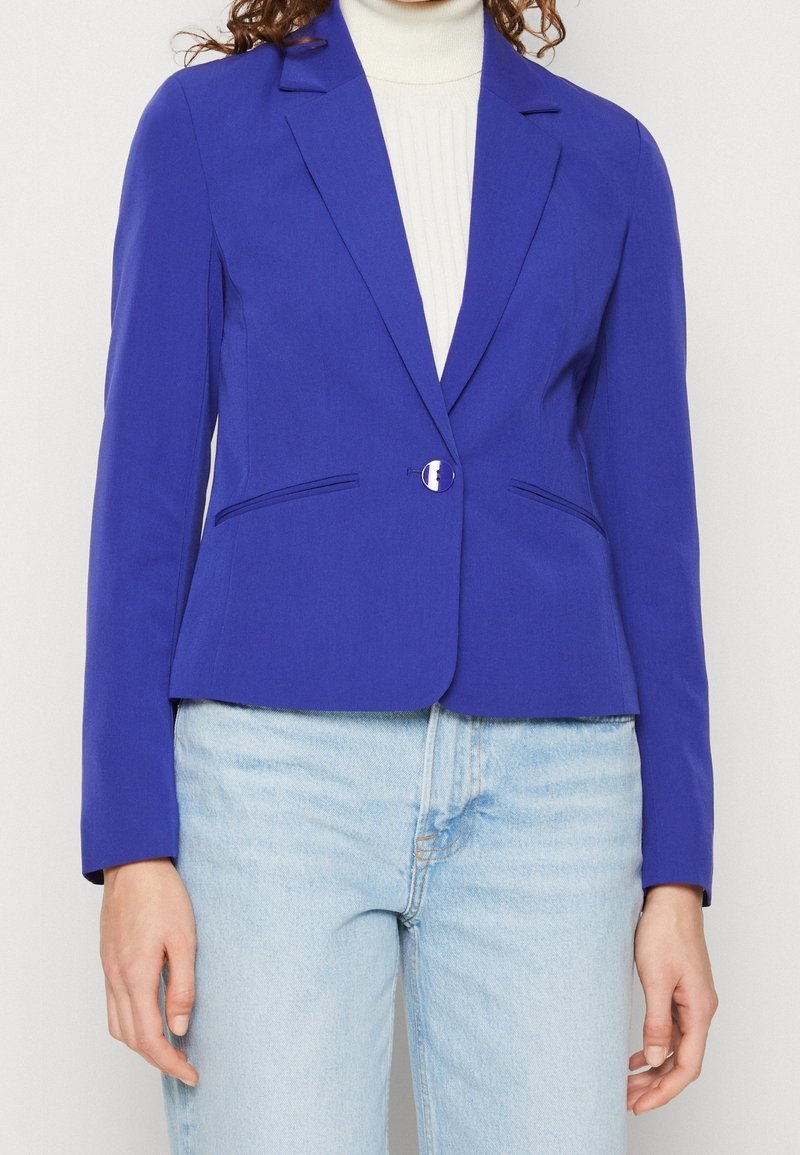 Femme portant un blazer bleu vif cintré avec un bouton sur un col roulé blanc et un jean bleu clair, montrant le haut du torse et les bras.