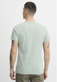 Camiseta de manga corta verde claro hecha de una tela suave, con cuello redondo y un ajuste relajado. Incluye tatuajes visibles en los brazos.