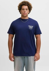 Mornarsko plava t-shirt kratkih rukava od pamuka. Ima bijeli otisak teksta na prsima i okrugli izrez. Nošena je s svijetlosivim hlačama.