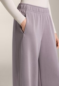 Světle fialové teplákové kalhoty vyrobené z měkké tkaniny, s volným střihem, elastickým pasem a bočními kapsami s žebrovanými akcenty po stranách.