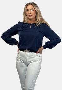 Granatowa bluzka z plisowanymi detalami i małym dekoltem w kształcie łezki, zestawiona z białymi dżinsami oraz czarnym paskiem z metalową klamrą.