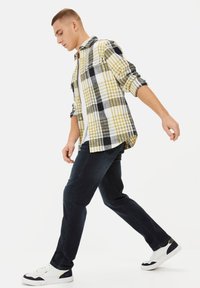 Geruite flanellen overhemd in geel, zwart en wit, open gedragen over een wit t-shirt. Gepaard met donkerblauwe spijkerbroek en witte sneakers.