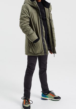 Olivengrøn polstret parka-jakke med hætte, der har lynlås og trykknapper, to forlommer og sorte ribstrikkede manchetter, båret over mørke bukser og farverige sneakers.