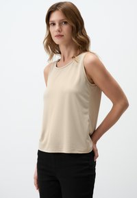 Top beige senza maniche realizzato in tessuto morbido, con scollatura rotonda e vestibilità comoda, abbinato a pantaloni neri.